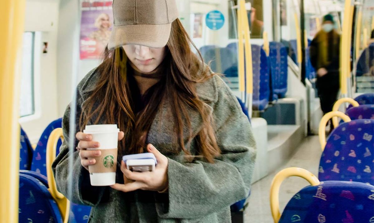 Girl holding a coffee, a phone and a charger standing in a train car 